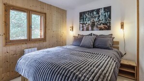 Un dormitorio con pared de paneles de madera, una cama grande con una colcha estampada, dos lámparas de noche y un cuadro con motivos de bosque.