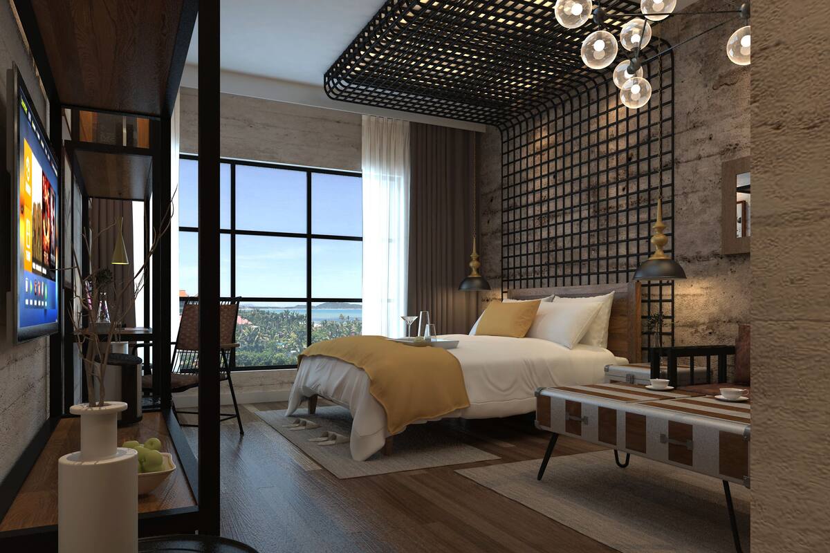 A modern bedroom with a large window, a bed with a yellow throw, a desk with a computer, and a unique ceiling design.