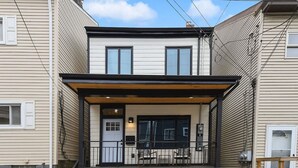 A two-story house with a front porch, a black door, and a small set of stairs leading to the entrance. - ★ HostWise Stays ★ Off-Street Parking, 2 Full Baths, Great Patio! (Pittsburgh)