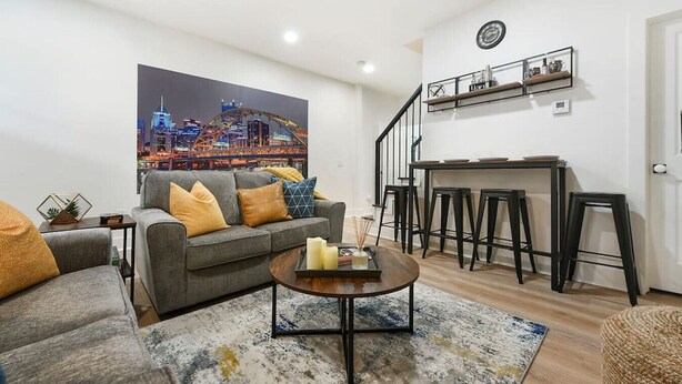 A living room with a sofa, coffee table, and a cityscape painting. There is a staircase leading to a kitchen area with stools. - ★ HostWise Stays ★ Off-Street Parking, 2 Full Baths, Great Patio! (Pittsburgh)