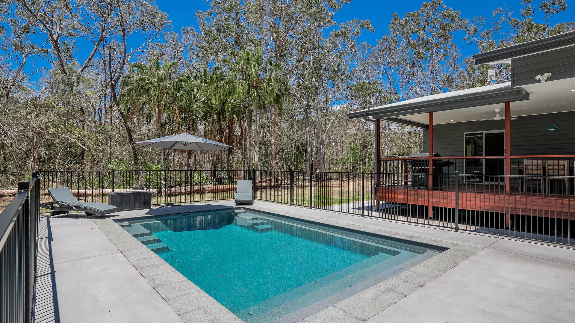A rectangular swimming pool with a clear blue water, surrounded by a concrete deck. There are two lounge chairs and a small table with an umbrella. The pool area is enclosed by a black metal fence, and there is a building with a red roof to the right.