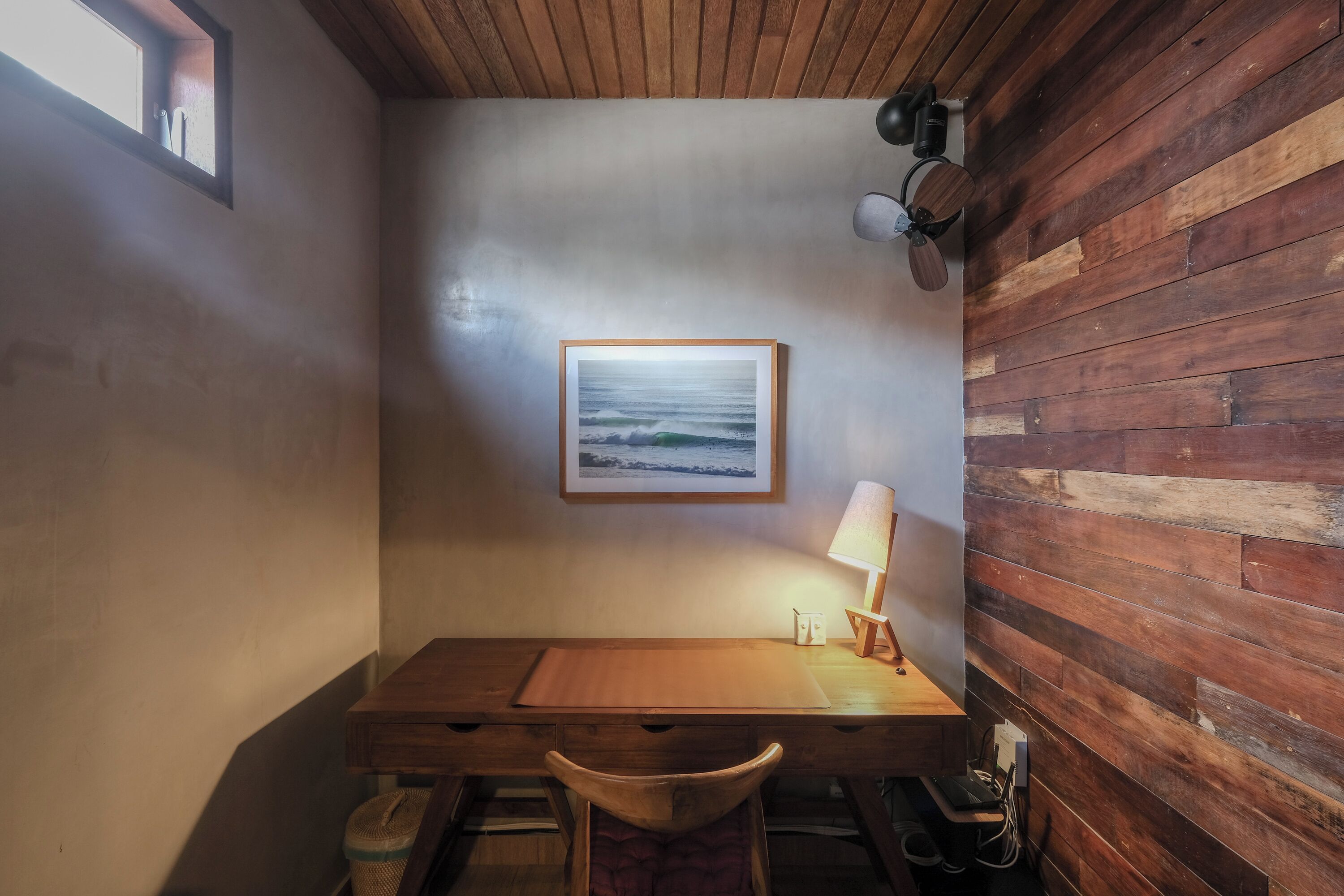 A corner room with a wooden desk, a chair, a framed picture of a beach, a lamp, and a ceiling fan.