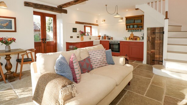 A cozy living room with a beige sofa, patterned cushions, and a woven throw blanket. A wooden dining table with chairs is visible in the foreground. The kitchen area is equipped with red cabinets, a stove, and a hanging pot rack. A staircase with wooden steps leads to an upper level. - The Little Hay Barn (Norwich)