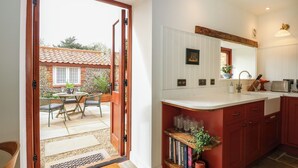 A kitchen with a wooden island, a dining area with outdoor seating, and a view of a stone house through an open door. - The Little Hay Barn (Norwich)