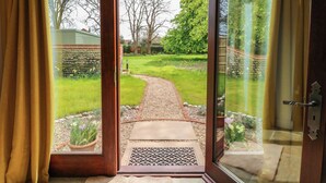 A glass door leading to a garden with a pathway, stone wall, and potted plants. - The Little Hay Barn (Norwich)