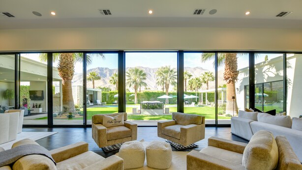 A spacious living area with large windows, beige sofas, and ottomans. - Mtn Views + Outdoor Oasis: Palm Springs Home! (Palm Springs)