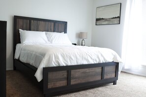 A bed with a dark wooden frame and white bedding, a bedside table with a lamp, and a framed picture on the wall. - In the Heart of Hobbs!  (Hobbs)