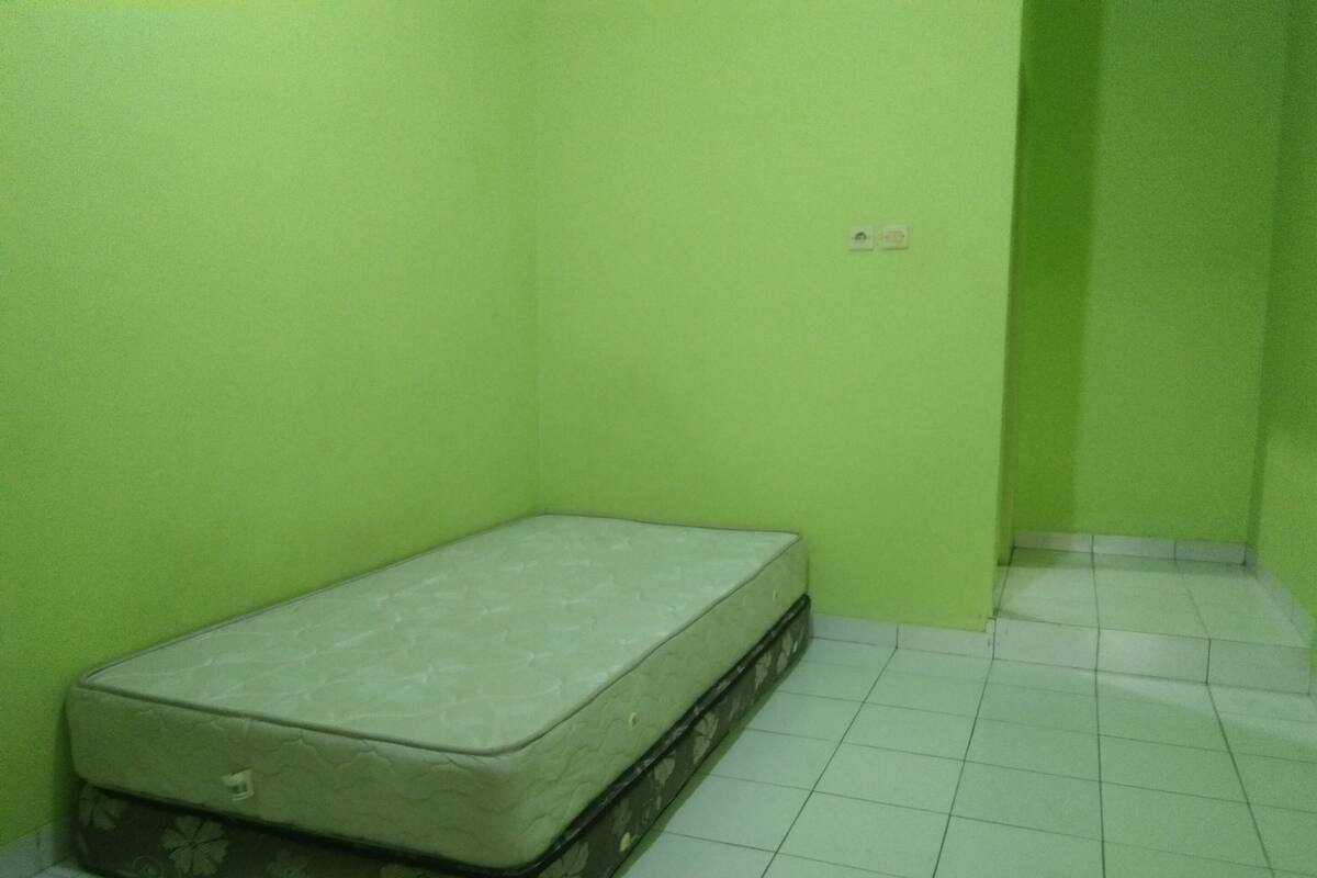A small, unadorned room with a mattress against a green wall and white tiled floor.