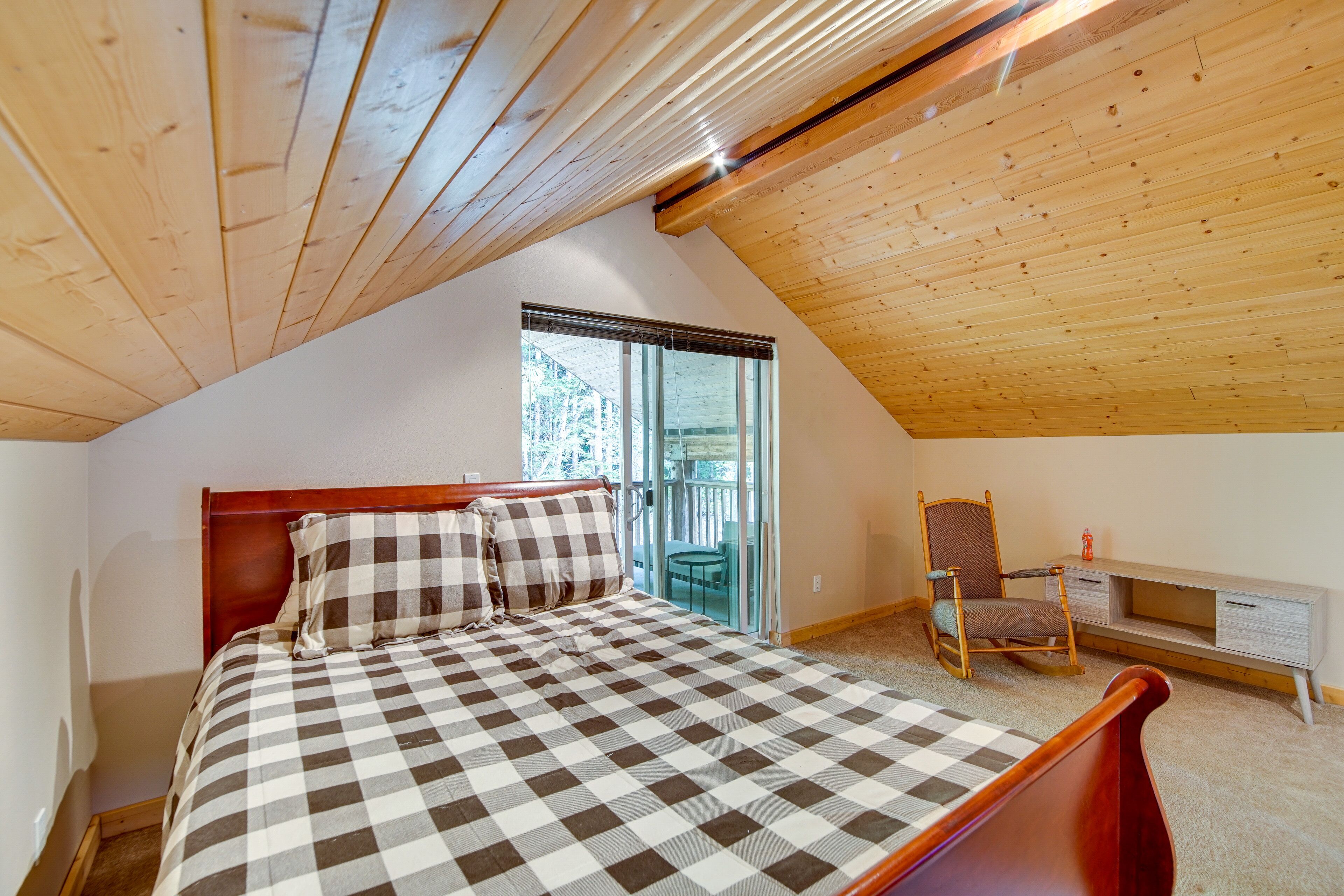 A bedroom with a checkered bedspread, a wooden ceiling, a chair, and a small table.