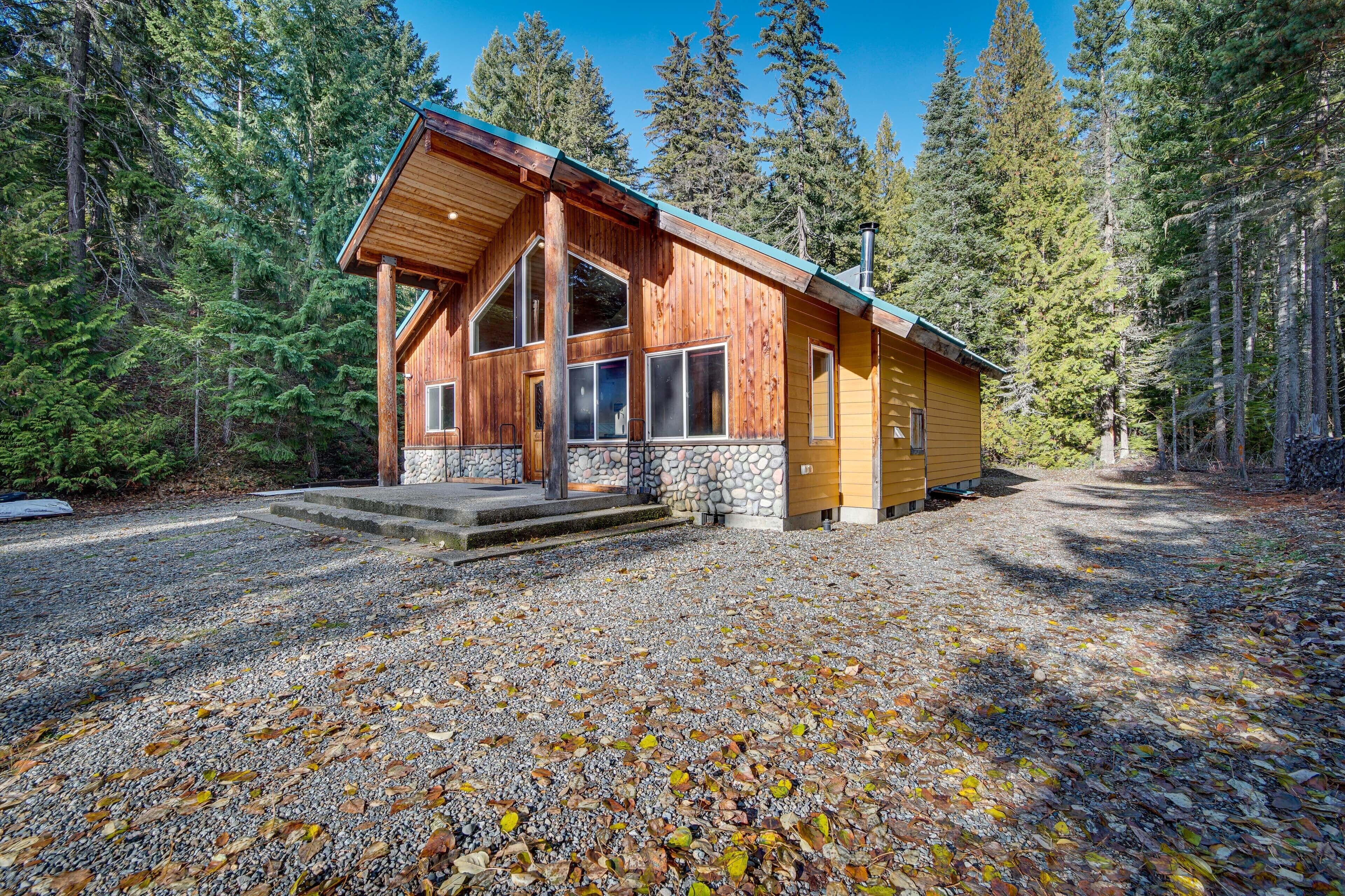 A wooden cabin with a stone foundation, large windows, and a gravel driveway surrounded by trees.