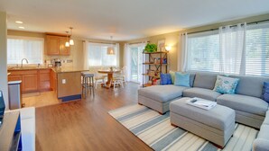 Ein modernes Wohnzimmer mit einem grauen Ecksofa, einem kleinen Hocker und einem Essbereich mit einem weißen Tisch und Stühlen.