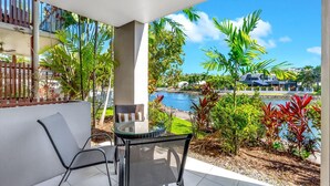 A balcony with a table and chairs, overlooking a body of water and a landscaped area with various plants.