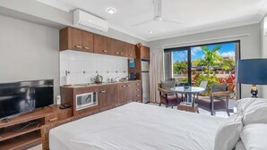 A hotel room with a bed, a TV, a kitchenette, and a balcony with a view of buildings and greenery.