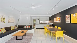 A modern living room with a sofa, coffee table, and yellow chairs. There is a kitchen area with a dining table and chairs. - Belle Escapes 73 Alamanda Palm Cove (Palm Cove)