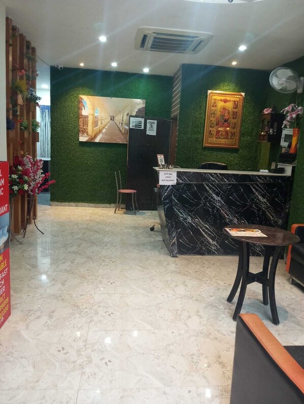 A hotel reception area with a marble floor, a reception desk, a chair, a small stool, a wall-mounted clock, and a framed picture. - Hotel Sangam Pacific And Restaurant (Dera Bassi)