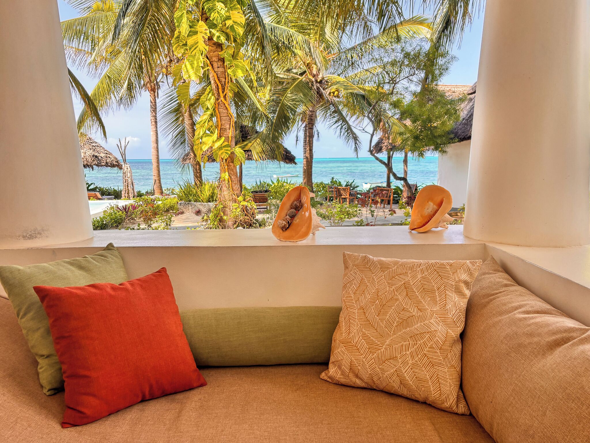 Una habitación con vista a una playa tropical, un alféizar con cocos y una zona de estar con cojines.