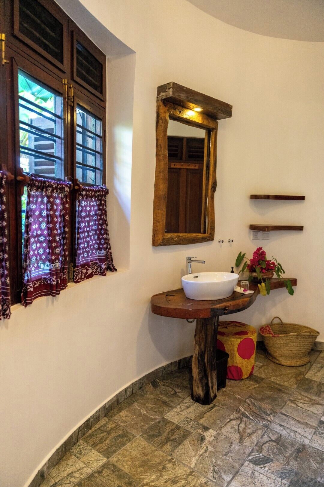 Un baño con tocador de madera, lavamanos blanco, espejo y ventana con cortinas estampadas.