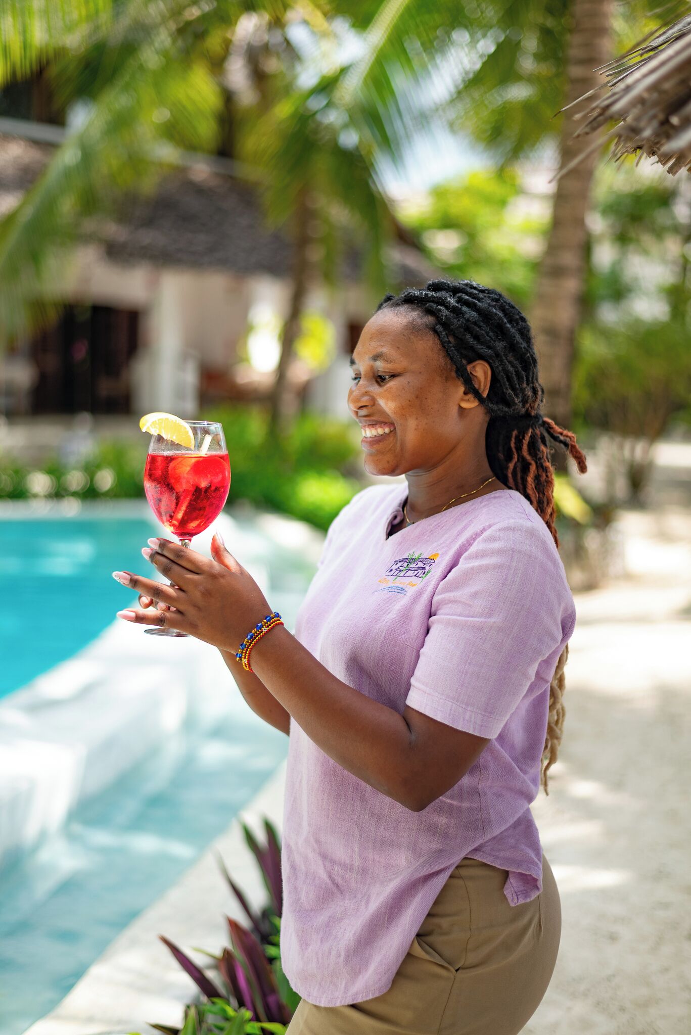 Una persona sostiene un vaso con una bebida roja, de pie junto a una piscina con un paisaje tropical.
