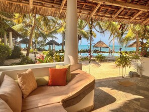 A room with a view of a beach and ocean, a cushioned seating area with pillows, and a thatched roof structure. - Villa Juliana (Jambiani)