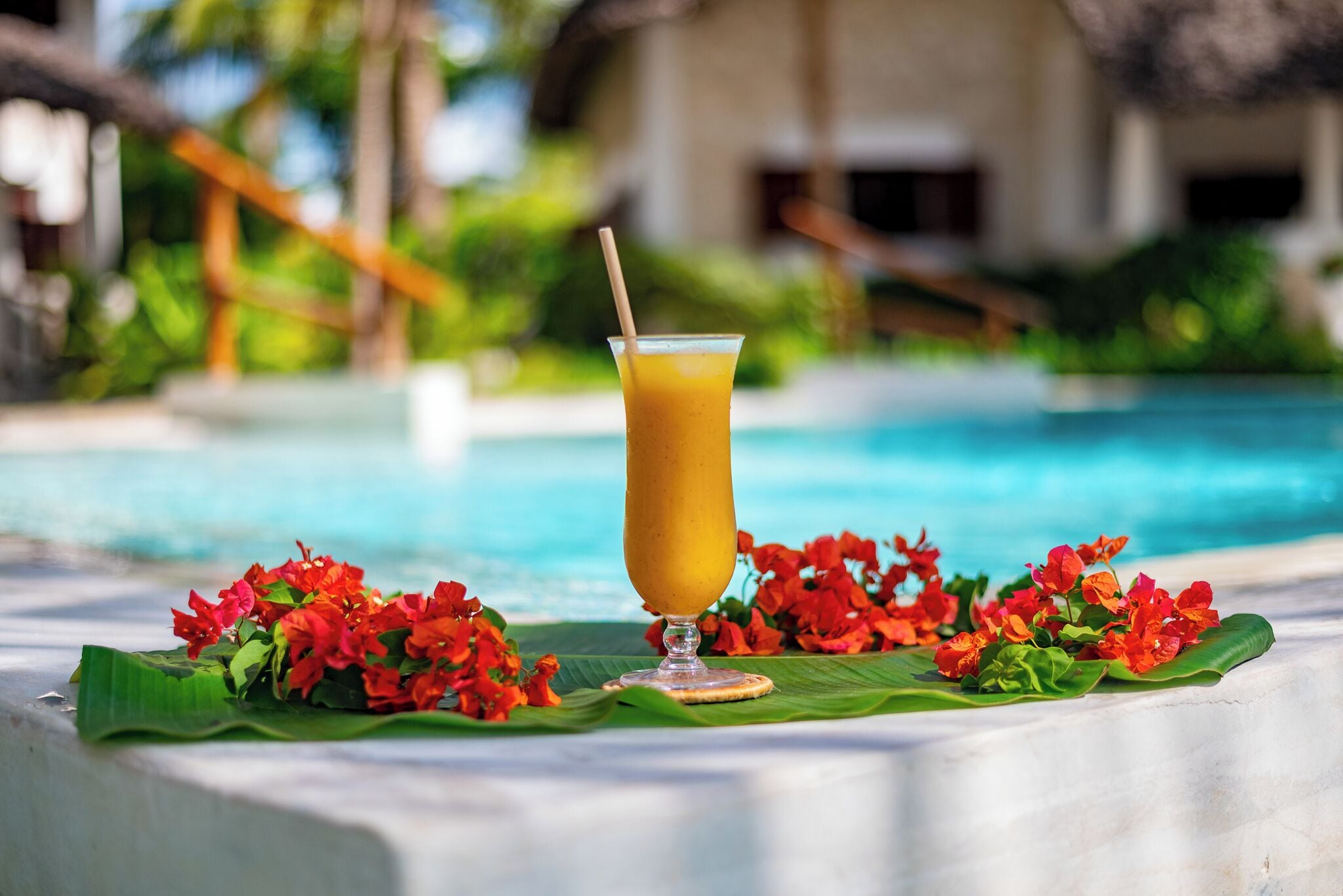 A clear glass of orange juice with a straw on a table by a pool with red flowers and a building in the background.