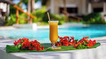 A clear glass of orange juice with a straw on a table by a pool with red flowers and a building in the background.