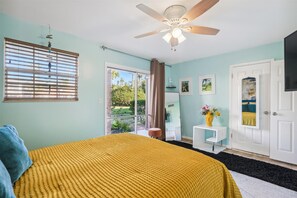 A bedroom with a bed, a ceiling fan, a window with blinds, a door, and a small table with a vase of flowers. - Chic Quarters; 15 min to Downtown!  (St. Augustine)