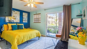 Ein Schlafzimmer mit Bett, Deckenventilator, Fenster mit Jalousien und Blick auf ein Gebäude.