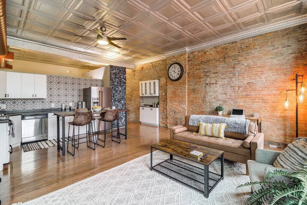 Una cocina moderna con armarios blancos, un comedor con mesa y sillas de madera, una sala de estar con sofá y mesa de centro, y una pared de ladrillo con un reloj.