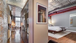 A modern hotel room with a brick wall, a large window, a bed, and a neon sign. - Modern Warehouse Loft | Walk to Rock Hall & Stadiums (Cleveland)