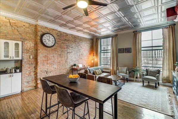 A spacious room with a brick wall, a dining table with chairs, a sofa, and a large window with curtains.