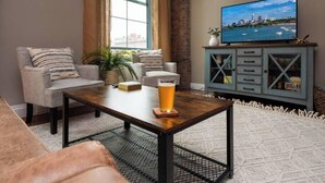 A living room with a sofa, a coffee table with a glass of beer, a television on a cabinet, and a window with curtains. - Modern Warehouse Loft | Walk to Rock Hall & Stadiums (Cleveland)