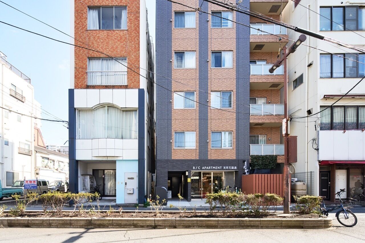 A multi-story apartment building with a B/C apartment sign, a visible entrance, and a small garden area in front.