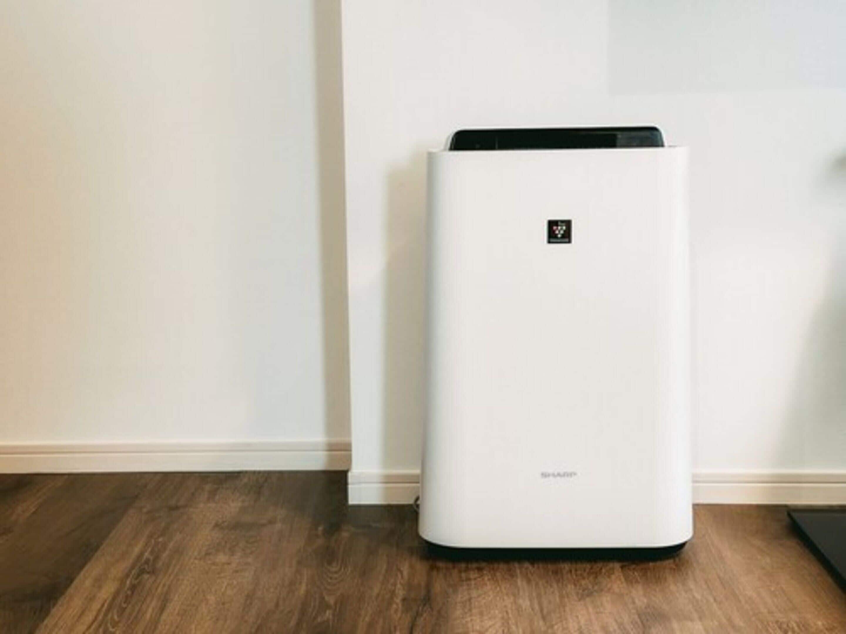 A white, modern air purifier with a control panel on the top, placed on a wooden floor against a plain wall.