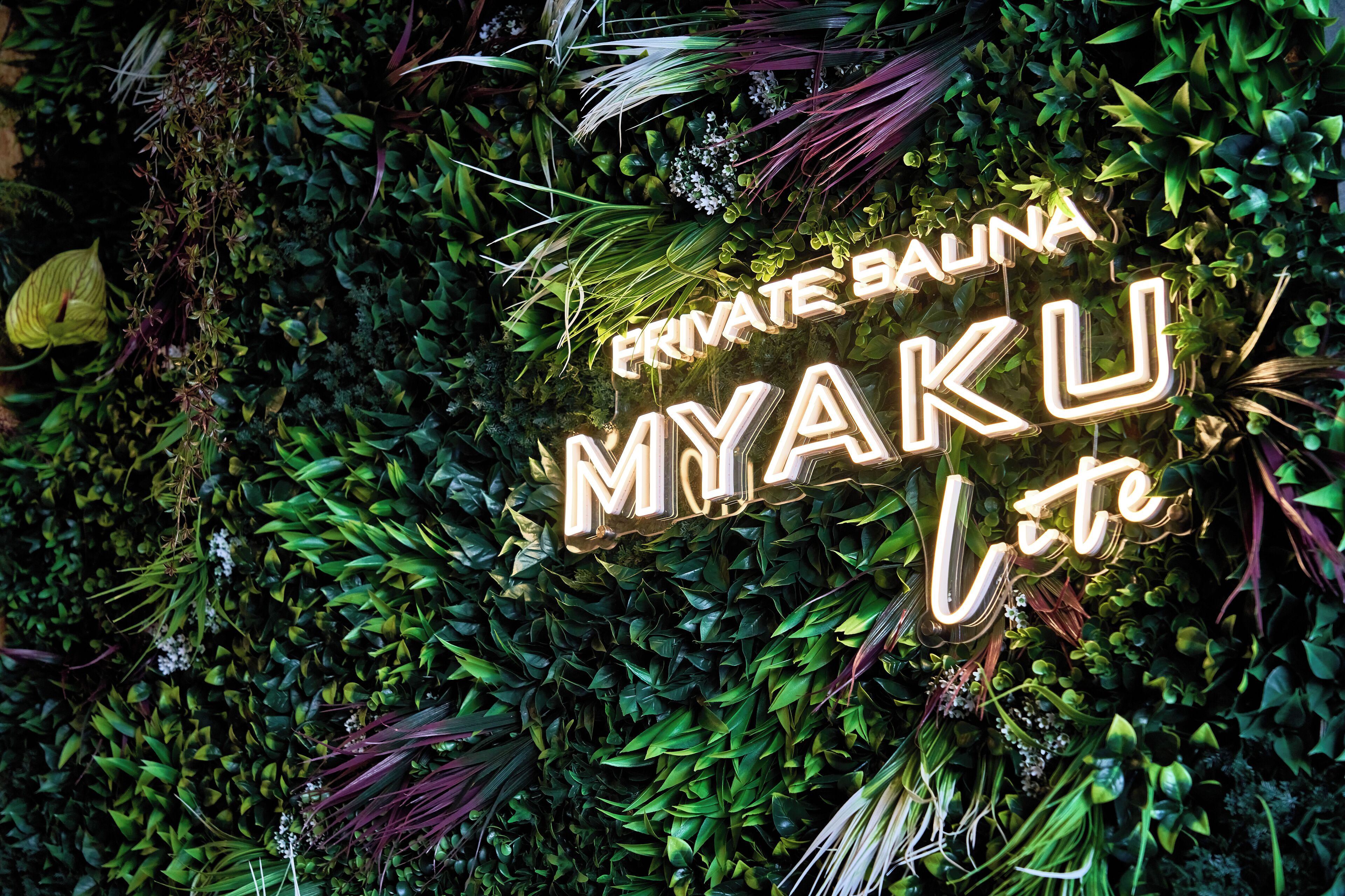 A neon sign with the text "PRIVATE BALINA MYAKU" against a backdrop of dense green foliage.