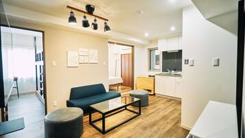 A modern living room with a sofa, ottoman, and coffee table, a kitchen area with cabinets and a TV, and a door leading to another room.