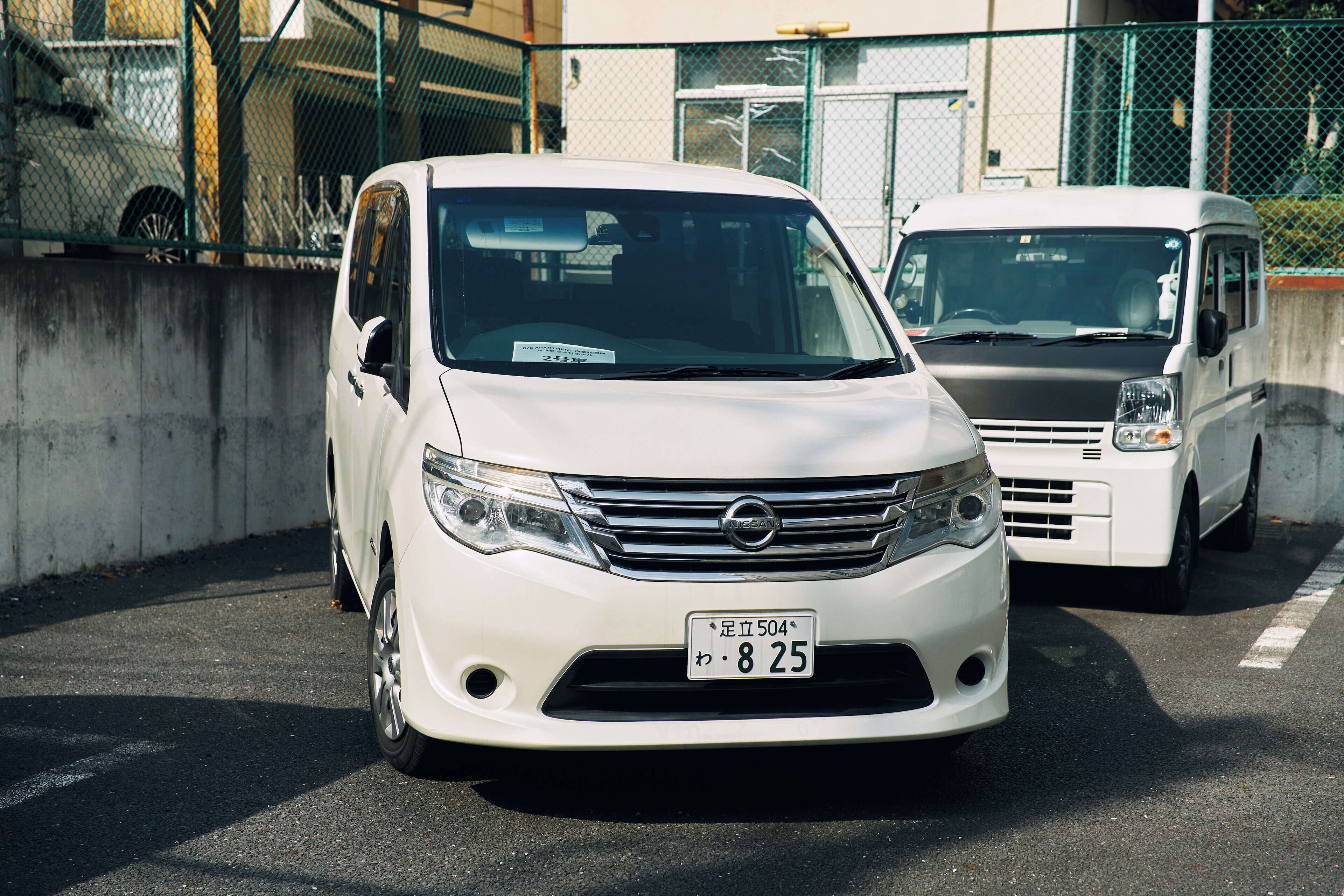 Una minivan blanca Nissan estacionada en un estacionamiento, con un muro de concreto y una cerca al fondo.