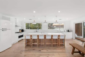 A modern kitchen with white cabinetry, a central island with bar stools, and a wooden bench. - Mogo MTB Base | Bike Workshop | Walk to Beach (Surf Beach)