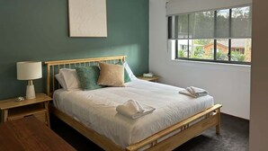 A bedroom with a bed, a nightstand, a lamp, a ceiling fan, and a window with a view. - Coastal Chillout at Mossy (Mossy Point)