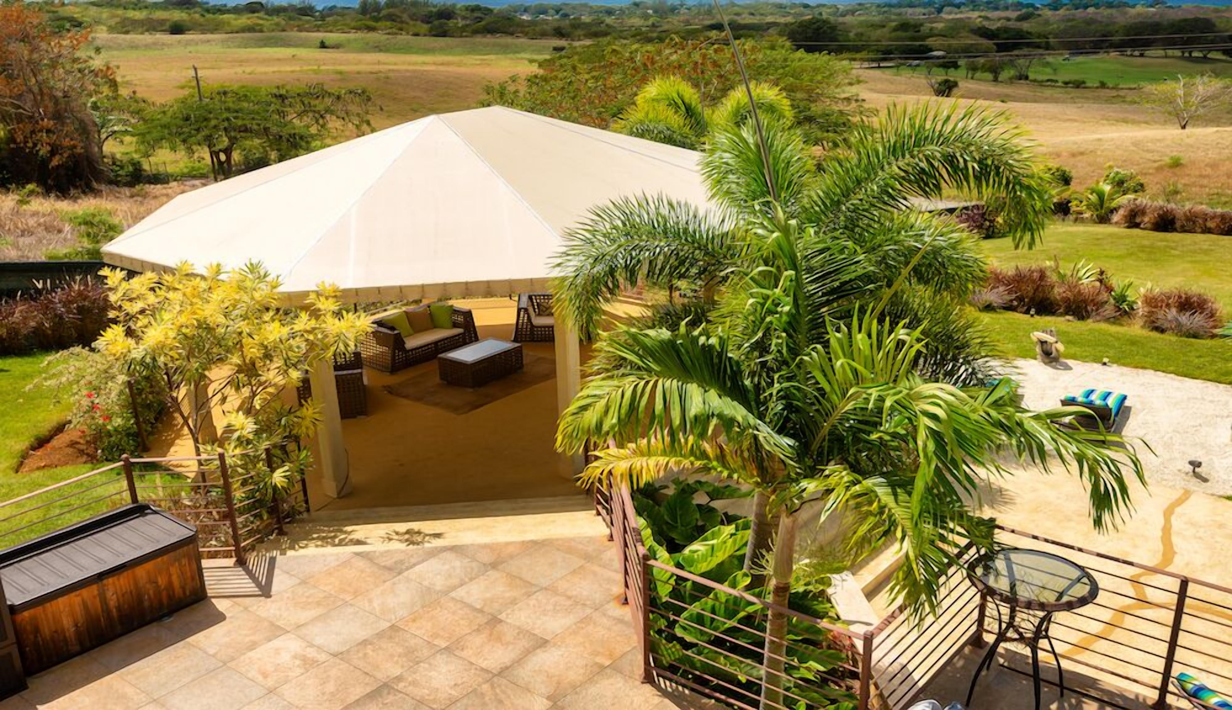 Un espace extérieur aménagé, abrité et doté de tables et de chaises, entouré d’une végétation luxuriante et offrant une vue sur la campagne.