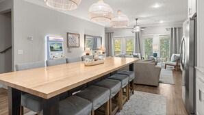 A modern kitchen with a long wooden table, grey upholstered benches, and pendant lighting. - Beatles Retreat, 5 min WALK to Beach + Golf Cart & Arcade Machine, Dog Friendly! (Miramar Beach)