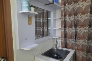 A small kitchen with a sink, a mirror, and a window. There are shelves above the sink and a curtain with a bird pattern on the window. - OYO Life 93160 Kost Jenny Karawang (Karawang)