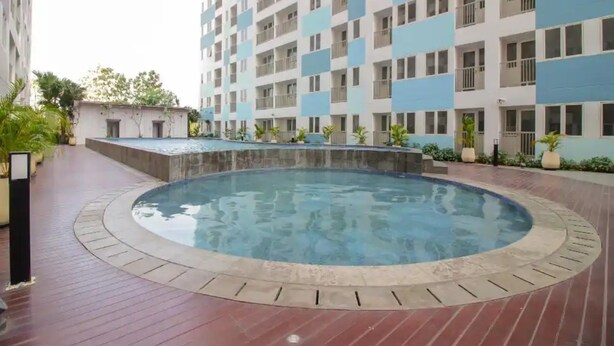 A swimming pool surrounded by a brick walkway, with a high-rise building in the background. - OYO Life 93139 Apartemen Grand Sentraland By Evi (Karawang)