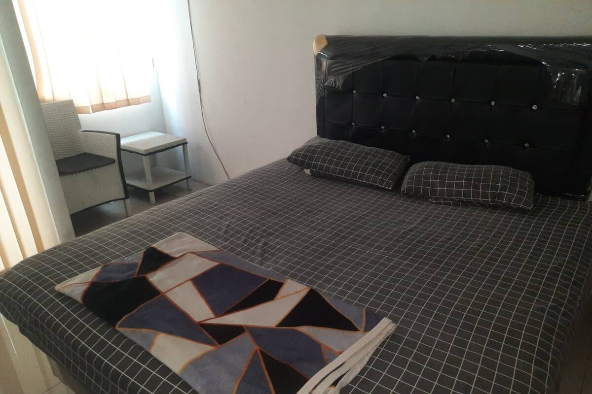A neatly made bed with a geometric-patterned blanket, a tufted headboard, and a small bedside table with a chair.