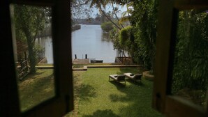 A view from a window of a grassy area with a water body in the background, a wooden deck, and a few lounge chairs. - Chittoor Kottaram - CGH Earth (Kanayannur)
