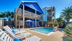 Un área junto a la piscina con sillones de descanso, una piscina y un edificio de dos pisos con terraza de madera.