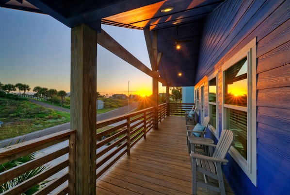 A wooden deck with a railing, overlooking a road and greenery, and a building with windows and a door.