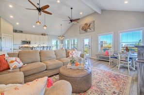 A spacious living area with a large sofa, ottoman, and dining table. There are ceiling fans, pendant lights, and a kitchen in the background.