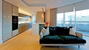 A modern living room with a dark green sofa, a kitchen in the background, and large windows offering a view of the city. - Modern Urban Retreat in Vibrant West London (London)