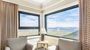Ein Eckzimmer mit zwei großen Fenstern und Blick auf die Berge, einem kleinen runden Tisch mit Lampe sowie zwei gepolsterten Sesseln.