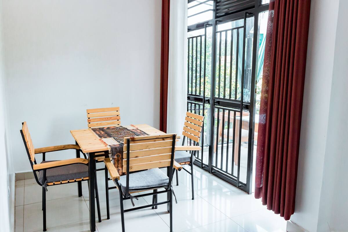 Un comedor con mesa y sillas de madera, un mantel decorado y vistas a través de un ventanal con cortinas rojas.